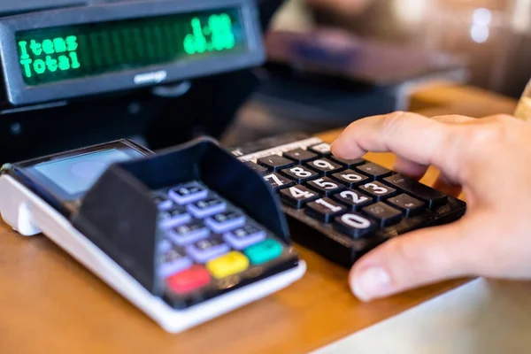 Selective focus to finger of women inserting password or pin code login on machine keypad of payment terminal. Cash desk with  payment terminal and blurry information screen in cafe.