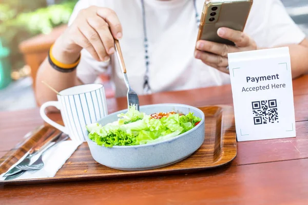Selective focus to Qr code payment tag. Blurry customer hand using smart phone to scan Qr code payment tag with coffee and salad on wooden table to accepted generate digital pay without money. Qr code
