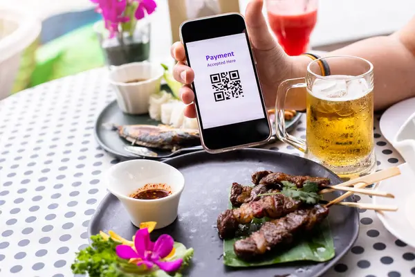 Hand holding smart phone to show scan QR code tag with blurry roast beef and beer in restaurant to accepted generate digital pay without money. Qr code payment concept.