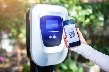 EV şöförü elektrik faturalarını ödemek için elektrikli araç şarj istasyonlarındaki QR kodunu taramak için akıllı telefonu kullanır. Para yerine QR tarama konsepti.