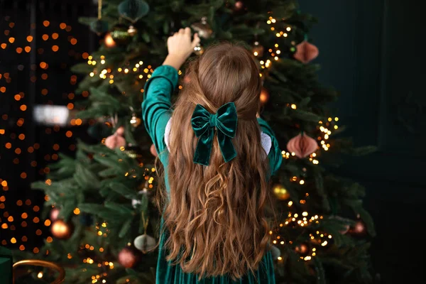 緑色のドレスを着た認識できない女の子と彼女の髪に赤いリボンの結び目でクリスマスツリーの後ろからリビングルームに立っています 後ろからの眺め 女の子はカメラに背を向けた — ストック写真
