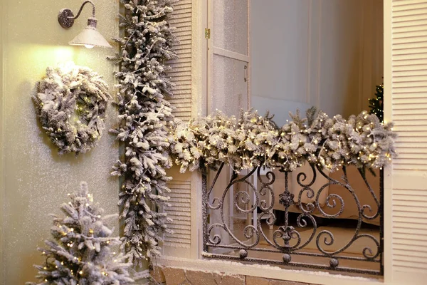 Old window with wrought-iron balcony decorated branches fir and glowing garland on Christmas. Snow covered balcony with wrought french iron railings with open shutters and doors.