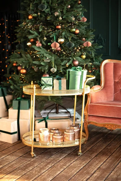 Christmas at home. New year winter home decor. Xmas tree with lights glowing garlands. Stylish loft living room interior with decorated Christmas tree, decorative trolley with gifts  and pink sofa.