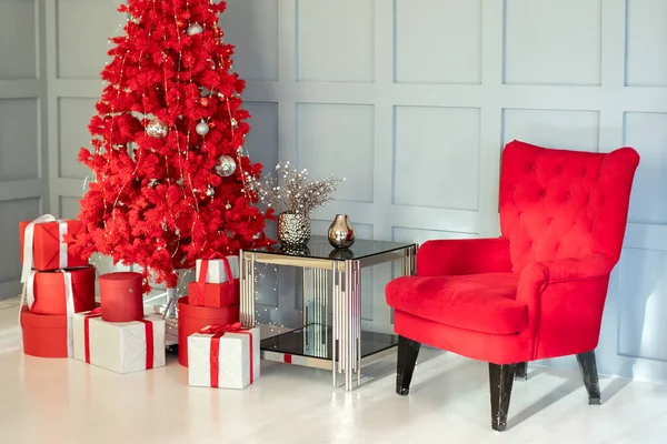 Stylish Christmas interior with elegant red armchair, coffee table and decorated Xmas tree. Modern Red Christmas tree with gifts in the living room. Happy new year interior background