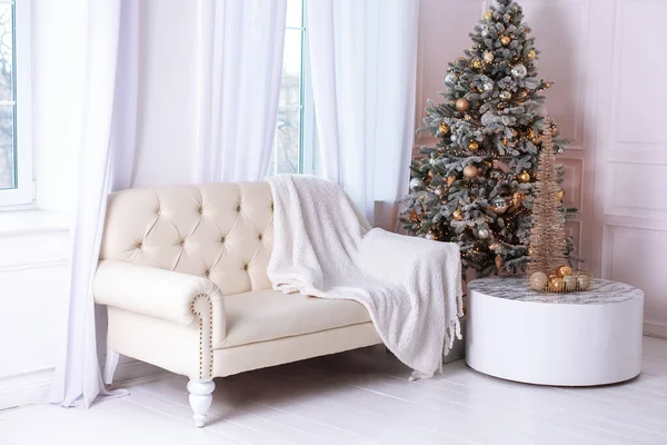 Christmas at home. New year winter home decor. Stylish white living room interior with decorated Xmas tree and sofa with plaid. A round mirror like sun hangs on wall