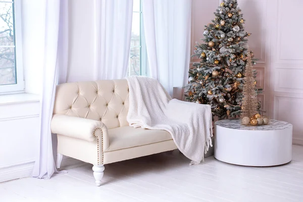 Christmas at home. New year winter home decor. Stylish white living room interior with decorated Xmas tree and sofa with plaid. A round mirror like sun hangs on wall