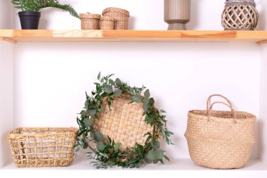 Boho interior of living room and wooden shelves with elegant decor, wicker basket and green houseplants in pots. Modern composition of wabi sabi interior with shelf with stylish home decoration. 