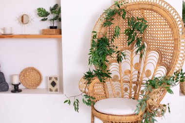 Design wicker chair and leaves decorations in stylish interior. Eco natural furniture. Rattan peacock armchair by white wall in living room. Wabi sabi room interior. Natural, rustic wedding decor.