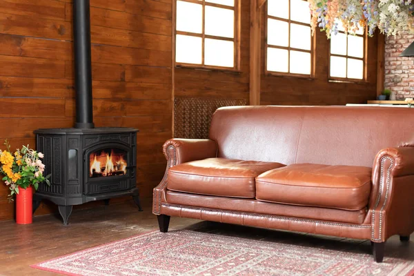 Interior cozy living room with comfortable brown leather sofa and carpet on floor. Black modern Cast iron wood stove at home. Traditional heating system. Classic fire place with wood burning inside