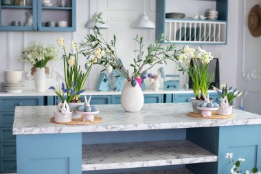Nergisler ve saksılarda bitkiler, ağaç dalları, vazoda paskalya yumurtası. Paskalya dekoru olan şık bir mutfak. Vazoda bir buket çiçek, seramik tabakta paskalya yumurtası ve sofrada çatal bıçak. 
