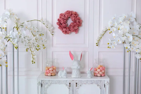 stock image Interior design of living room interior with wooden dresser, stylish easter bunny sculpture and eggs. Minimalist composition with white easter rabbits ceramic statuette and many colorful eggs in vase