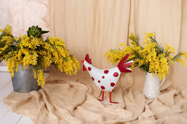 Easter home interior. Spring flowers in a vase and statuette easter chicken on a light beige background. Bouquet mimosa yellow flowers in vase. Branches yellow flowers in a bouquet. Mother\'s day.