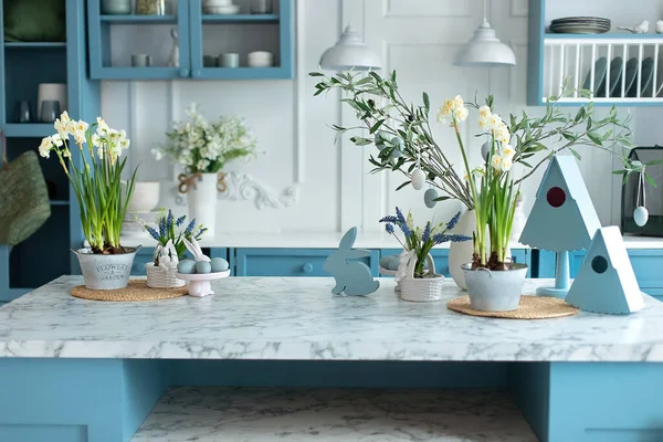 Home interior with easter decor. Kitchen utensils on table. Blue kitchen interior with furniture. Stylish cuisine with flowers in vase. Daffodils in pot and branches tree in vase with easter eggs.