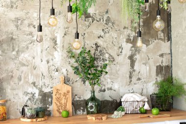 Duvardaki sert çimento alçısının arka planındaki tavan arası lambaları. Dahili Scandi açık mutfak. Ahşap raflar, çeşitli yemek takımları, saksılarda bitkiler ve mutfakta süslemeler.. 