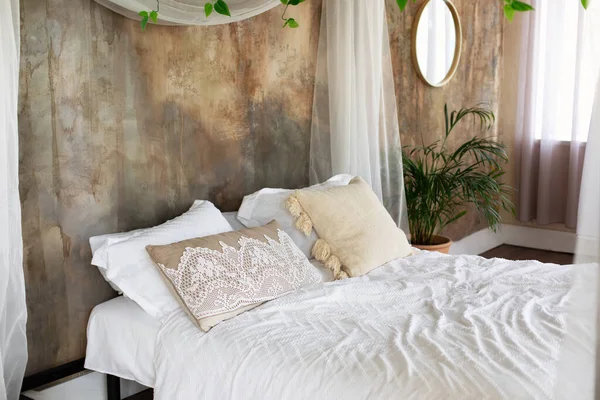Soft different pillows, blanket and duvet cover on bed. Boho style Bedroom with bedding. Interior cozy bedroom in loft style bedroom with bed decorated flowing white curtains and plant in pot.