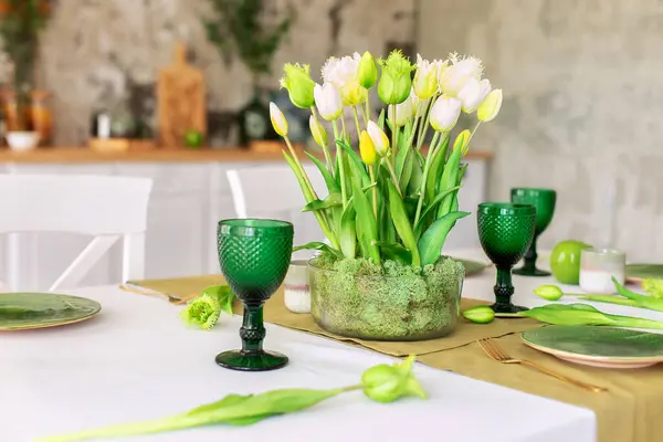Table festive setting with tulips flowers and stabilization moss in glass vase. Wedding table decoration. Beautiful served table with runner on table, wineglass and plates in living room