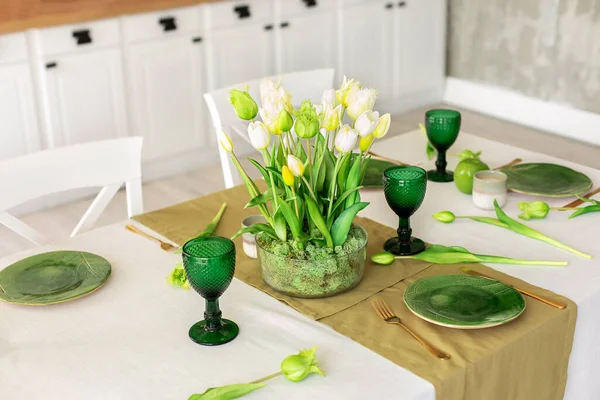 Table festive setting with tulips flowers and stabilization moss in glass vase. Wedding table decoration. Beautiful served table with runner on table, wineglass and plates in living room