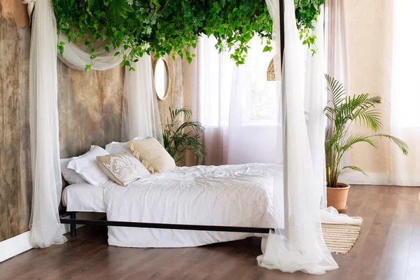 Cozy light bedroom in Scandinavian style bedroom, canopy bed, plant in pot. Bed decorated flowing white curtains canopies. Interior boho design bedroom with composition from garland flowers and plants