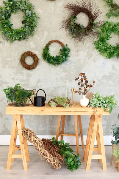 Interior workplace florist. Wooden table on which there are green plants in flowerpots, watering can, flowers on a background of a grey wall. Living room in rustic style in summer. Flowers shop.