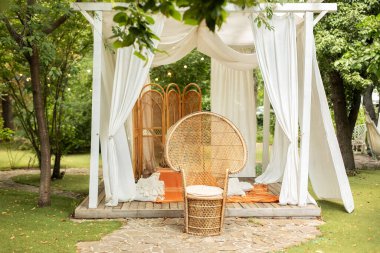 Rattan tavus kuşu koltuğu ve dekoratif katlama ekran ayırıcısı. Beyaz perdeleri akan yaz çardağı. Düğün boho dekorasyonu. Hasır mobilyalarla dışarıdaki terası dekore et. Arbour 'un açık hava tasarımı