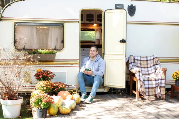 Rest man sitting on the doorstep of the motor home holding iron mug. Smiling handsome man sitting on the porch of his RV trailer holding a mug of coffee. Campsite on caravan or camper van in forest.