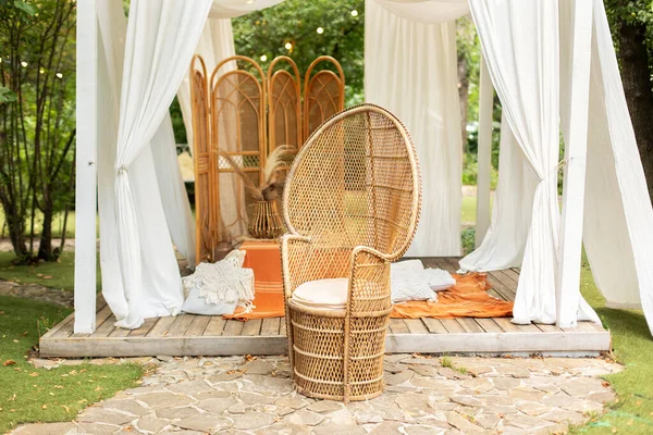 Rattan peacock armchair and decorative folding screen divider. Summer gazebo with flowing white curtains. Wedding boho decoration. Decor outdoor terrace with wicker furniture. Outdoor design of arbour