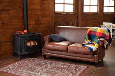 Interior cozy living room with wood burning stove inside, comfortable sofa, rustic wooden walls and carpet on floor. Black modern Cast iron wood stove at home. Fragment of interior of country house. 