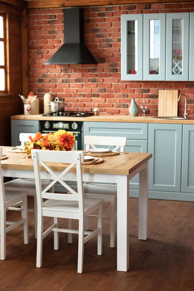 Interior design scandinavian kitchen with utensils, dishes, plates. Stylish dining room with wooden table and chairs. Cozy cuisine decorated with fall decor and table setting flowers and pumpkins.