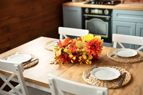 Cozy table setting with fall decor, flowers and utensils in dining room. Bouquet of fall flowers. Cozy kitchen interior. Festive table with wicker napkins, white plates and cutlery on wooden table.