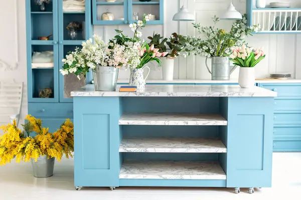 Blue kitchen interior with furniture. Stylish cuisine with flowers in vase. Wooden kitchen in spring decor. Cozy home decor. Kitchen island in dining room. Kitchen utensil, dishes and plate on shelves
