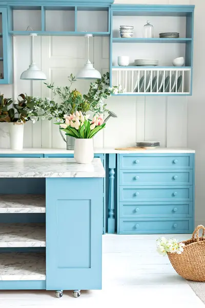 Kitchen island in dining room. Stylish cozy cuisine with tulips flowers in vase. Wooden kitchen in spring decor. Kitchen utensil, dishes and plate on shelves. Blue kitchen interior with furniture