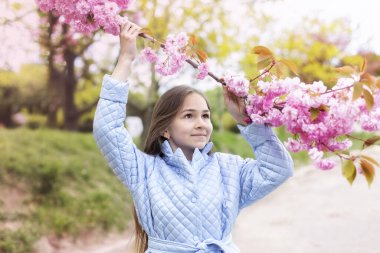Trençkotlu gülümseyen kız çiçek açan sakura bahçesinde gülümsüyor ve hayaller kuruyor. Açık hava moda portresi. Çiçek açan bahar parkında şık mavi trençkot giyen küçük kız.. 