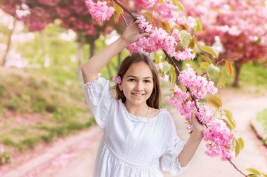 Sakura bahçesindeki çiçek açan ağaçta beyaz elbiseli, gülümseyen genç kız. Kadın güzelliği ve doğa konsepti. Çiçekli bahar parkında yürüyen beyaz elbiseli güzel bir kızın portresi.. 