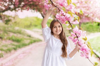 Sakura bahçesindeki çiçek açan ağaçta beyaz elbiseli, gülümseyen genç kız. Kadın güzelliği ve doğa konsepti. Çiçekli bahar parkında yürüyen beyaz elbiseli güzel bir kızın portresi.. 