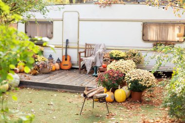 Karavanın yanındaki koltukta. Bahçe mobilyaları olan bir karavan evi verandası. Bahçesinde ateş püsküren bir kamp alanı. Sonbahar çiçekleri saksısı ve Cadılar Bayramı için süslenmiş balkabaklarıyla iç bahçe kamp alanı.