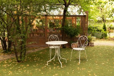 Bahçe mobilyalarının olduğu arka bahçe. Evdeki rahat teras. Beyaz demir sandalyeler ve dışarıda ya da dışarıda dinlenmek için bir masa. Bahçede sonbahar atmosferi - masa, sandalyeler ve düşmüş sarı yapraklar. 
