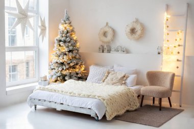 Konforlu İskandinav beyaz iç tasarım yatak odası Noel ağacı, yatak ve yastıklar, bej koltuk ve çelenkli dekoratif merdiven. Pencerede beyaz Noel yıldızları var. Bir lamba yıldızı