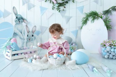 Bahar tarzında çocuk odasında küçük bir kız. Kıvırcık saçlı, pembe elbiseli, paskalya süslemeli tatlı bir kız bebek. Paskalya yumurtaları ve dekoratif figürlü tavşanları olan mutlu çocuk.