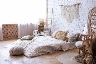 Boho tarzı iç dizaynı olan rahat bir yatak odası, yerde yatak yatağı, yastıklar, ev dekorasyonu, tavus kuşu koltuğu, merdiven, beyaz tuğla duvara karşı makrome panel. Klas ev mi, daire mi kavramı.