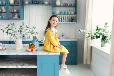 Paskalya pastası, çiçekler, paskalya yumurtaları ve mutfaktaki masada oturan sarı elbiseli, gülümseyen küçük sarışın kız. Mutlu çocuk. Kız evde paskalya yumurtası avlıyor.