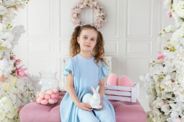 Mavi elbiseli, elinde oyuncak paskalya tavşanıyla güzel bir kız. Tatlı küçük kız Paskalya yumurtalarını kovalıyor. Paskalya yumurtası avla. Paskalya iç tasarım odası bahar dekorasyonu. Çiçekler evde kemerli.