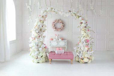 Beyaz oturma odasının içi pembe koltuk puf ve dekoratif çiçek kemeriyle dolu. Tatil bahar dekoru. Duvarında çelenk, çekmece, seramik tavşan ve paskalya yumurtası olan Paskalya bayramı dekorasyonu.