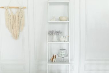 Duvarda paskalya süslemeleri, şenlik çelenkleri, seramik tabaklar ve çatal bıçaklarla ahşap bir raf var. Paskalya oturma odasının rahat iç tasarım kompozisyonu. İçi tavşan heykeli ve eastrer yumurtalarıyla dolu.. 