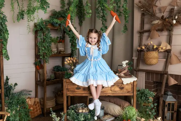 Mavi elbiseli güzel küçük kız, elinde havuçlarla Paskalya 'nın arka planındaki iç odasının havuçlarını tutuyor. Çocuklar için sağlıklı yiyecekler. organik yiyecekler, vejetaryen, sebzeler