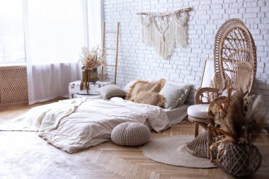 Wabi-sabi. Boho tarzı iç dizaynı olan rahat bir yatak odası, yerde yatak yatağı, yastıklar, ev dekorasyonu, tavus kuşu koltuğu, merdiven, beyaz tuğla duvara karşı makrome panel. Modern ev kavramı