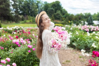 Yakından bakınca, beyaz elbiseli güzel bir kız ve şakayık tarlasında çiçeklerle yürüyen hasır bir şapka. Gülümseyen mutlu kız çiçekleri tutuyor. Çiçek açan bahçede pembe şakayıklar.