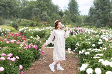 Beyaz elbiseli güzel bir genç kız portresi ve şakayık tarlasında çiçeklerle yürüyen hasır şapka. Gülümseyen mutlu kız çiçek tutan pembe şakayık çiçekli bahçede