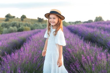 Hasır kayıkçı şapkalı ve beyaz elbiseli güzel bir kız sahada lavanta topluyor. Portre şirin, mutlu kız çocuğu lavanta çalılarının ortasında duruyor. Provence. Çocuklar yazları açık havada rüya görür.