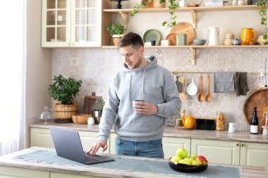 Gülümseyen adam elinde fincan kahveyle mutfak masasında duruyor dizüstü bilgisayarında e-posta mesajı okuyor. Evden çalışan serbest çalışan erkek uzaktan kumanda. Genç adam bilgisayarını kullanarak evdeki arkadaşlarıyla sohbet ediyor.. 