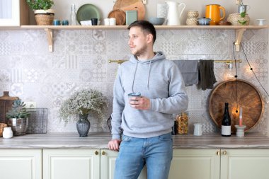 Sabahları mutfakta gülümseyen ve kahve içen yakışıklı bir adam. Bir fincan çayla kameraya bakan bir adam. Tişörtlü ve kot pantolonlu genç adam evde rahat duruyor. Yaşam tarzı kavramı.